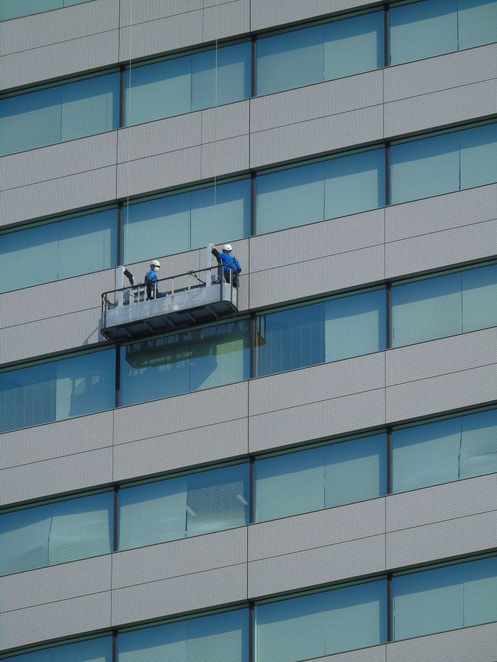 Deux ouvriers nettoient les vitres d’un immeuble moderne en verre à l’aide d’une plateforme suspendue.